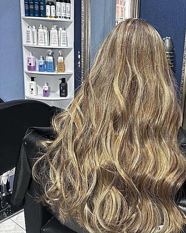 Balayage rubio miel en ondas largas, de espaldas en un sillón del salón, con la repisa de productos al fondo
