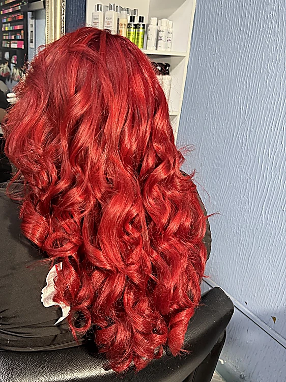 Cabello rojo intenso en rizos grandes, de espaldas en el sillón, con repisa de productos al fondo
